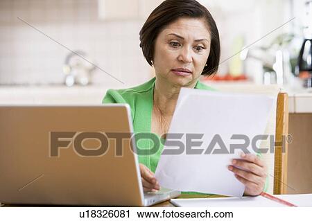Stock Image - Woman in kitchen with laptop and paperwork looking worried. Fotosearch