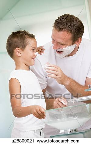 Stock Image - Man in bathroom putting shaving cream on young boy's nose. Fotosearch