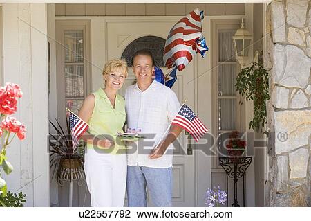 Stock Image - Couple at front door on fourth of July with flags and cookies smiling. Fotosearch