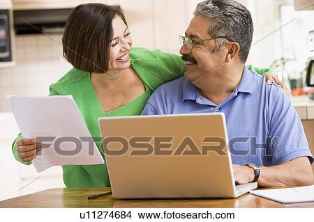 Picture - Couple in kitchen with laptop and paperwork smiling. Fotosearch