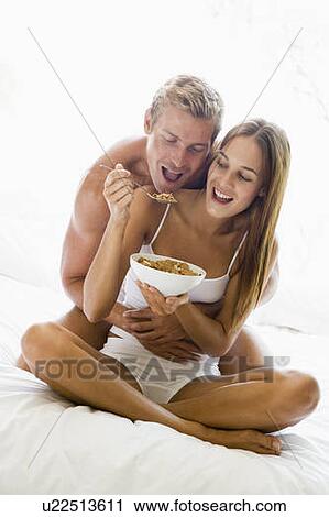 Stock Image - Couple sitting in bed eating cereal and smiling. Fotosearch