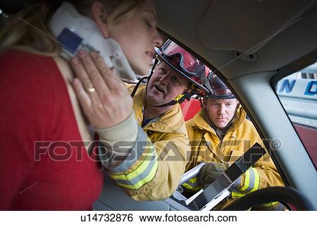 Stock Photograph - Fireman helping woman with neck brace while another fireman uses the jaws of life on a car door (selective focus). Fotosearch