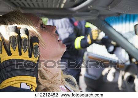 Stock Image - Injured woman in car with firefighter in background cutting out windshield (selective focus). Fotosearch