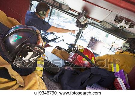 Stock Image - Two firefighters driving a fire engine with gear in the back (selective focus). Fotosearch
