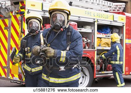 Stock Photograph - Two firefighters with hose and axe walking away from fire engine and another firefighter in background (selective focus). Fotosearch