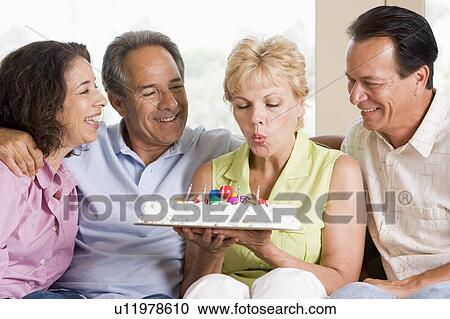Stock Image - Two couples in living room smiling with woman blowing out candles on cake. Fotosearch
