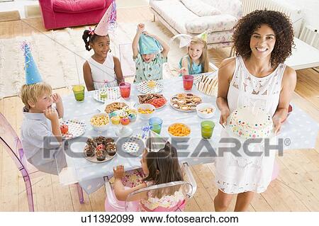 Stock Photo - Young children at party sitting at table with mother carrying cake and smiling. Fotosearch