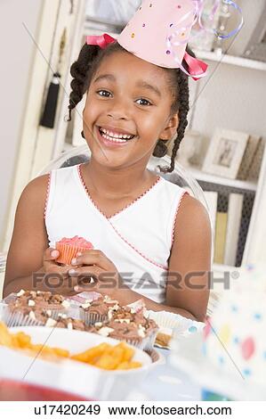Stock Photo - Young girl at party sitting at table with a cupcake smiling. Fotosearch