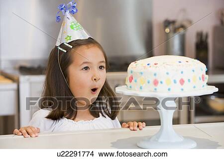 Young girl wearing party hat at kitchen counter looking at cake smiling View Large Photo Image Picture - Young girl wearing party hat at kitchen counter looking at cake smiling. Fotosearch
