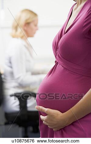 Stock Photograph - Pregnant woman at work holding belly with coworker in background. Fotosearch