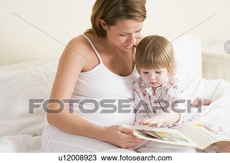 Stock Image - Pregnant woman in bedroom reading book with daughter. Fotosearch