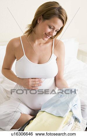 Stock Photograph - Pregnant woman packing baby clothing in suitcase smiling. Fotosearch