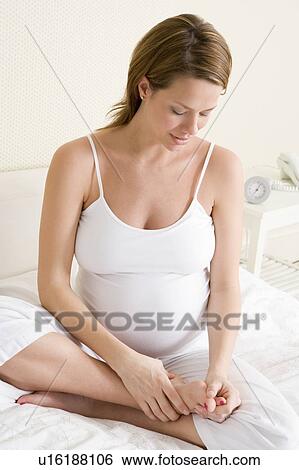 Stock Photograph - Pregnant woman sitting in bed rubbing feet. Fotosearch