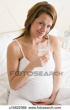 Stock Photo - Pregnant woman sitting in bedroom with glass of water smiling. Fotosearch