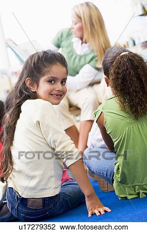 Stock Image - Student in class looking at camera with teacher reading in background (selective focus). Fotosearch