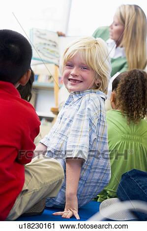 Stock Image - Student in class looking at camera with teacher reading in background (selective focus). Fotosearch