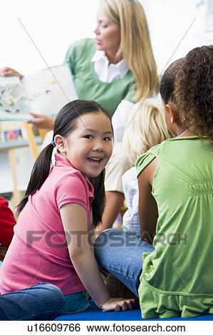 Stock Photograph - Student in class looking at camera with teacher reading in background (selective focus). Fotosearch