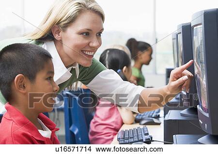 Picture - Teacher helping student at computer terminal with students in background (selective focus/high key). Fotosearch