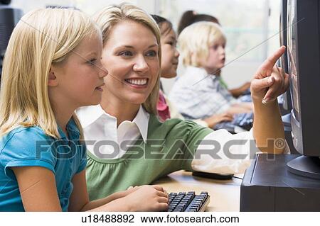 Stock Image - Teacher helping student at computer terminal with students in background (depth of field/high key). Fotosearch