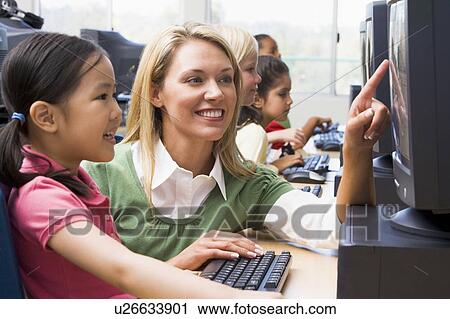 Stock Image - Teacher helping student at computer terminal with students in background (depth of field/high key). Fotosearch