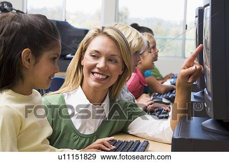Stock Photo - Teacher helping student at computer terminal with students in background (depth of field/high key). Fotosearch