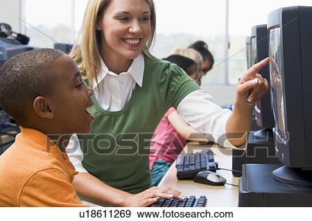 Stock Photo - Teacher helping student at computer terminal with students in background (selective focus/high key). Fotosearch