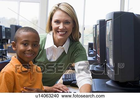 Stock Image - Teacher sitting with student at computer terminal with students in background (selective focus/high key). Fotosearch