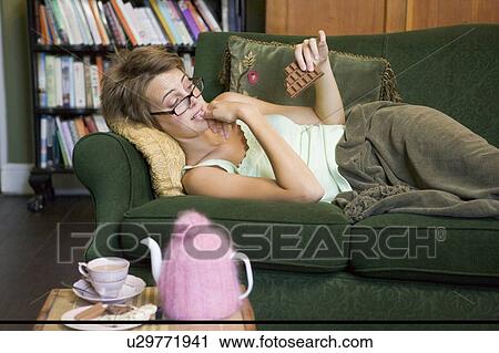 Stock Image - Young woman lying on sofa at home eating chocolate. Fotosearch