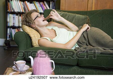 Stock Photo - Young woman lying on sofa at home eating chocolate. Fotosearch