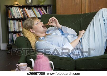 Stock Photography - Young woman lying on sofa at home eating chocolate. Fotosearch