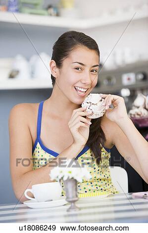 Young woman sitting at a table eating a sweet treat View Large Photo Image Stock Photo - Young woman sitting at a table eating a sweet treat. Fotosearch