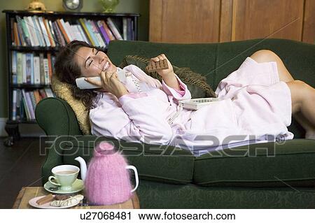 Stock Image - Young woman lying on sofa at home eating chocolate and talking on telephone. Fotosearch