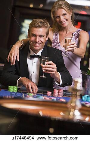 Picture - Couple in casino at roulette table holding up champagne and smiling (selective focus). Fotosearch