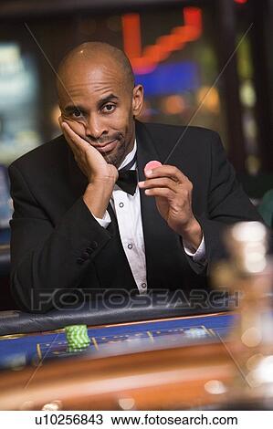 Stock Image - Man in casino playing roulette and holding chip (selective focus). Fotosearch