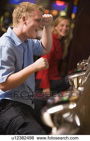 Stock Photo - Man in casino playing slot machine smiling with people in background (selective focus). Fotosearch