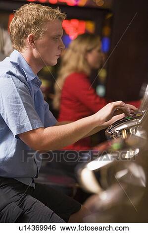 Stock Photograph - Man in casino playing slot machine with people in background (selective focus). Fotosearch