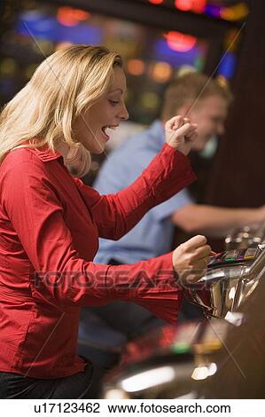 Stock Image - Woman in casino excited playing slot machine with people in background (selective focus). Fotosearch