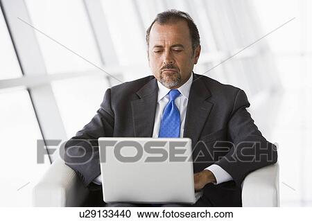 Stock Image - Businessman sitting indoors with laptop (high key/selective focus). Fotosearch