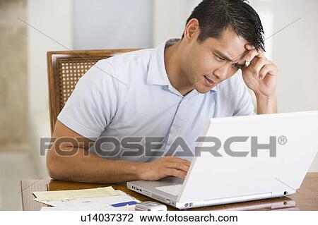 Stock Image - Man in dining room using laptop and frowning. Fotosearch