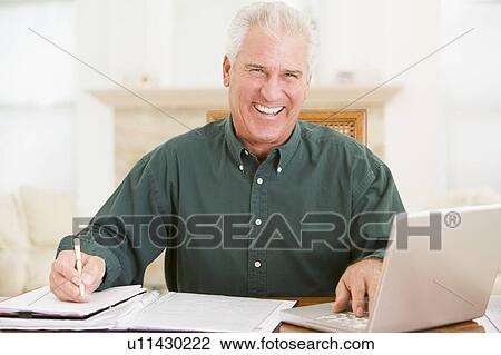 Stock Image - Man in dining room with laptop and paperwork smiling. Fotosearch