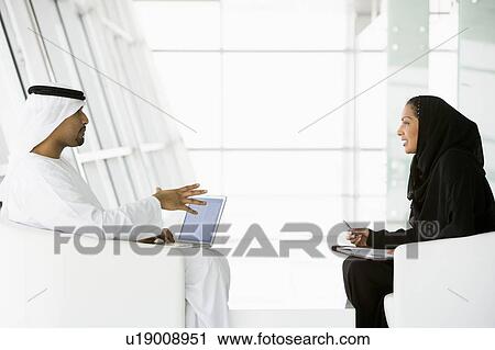 Stock Image - Two businesspeople indoors with a laptop talking and smiling (high key/selective focus). Fotosearch