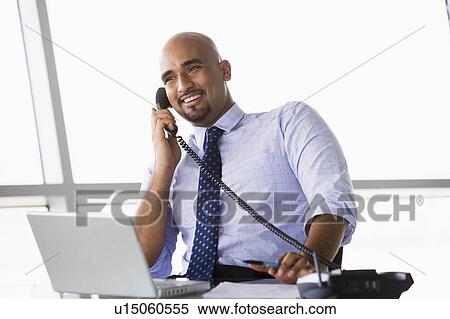 Stock Photography - Businessman in office on telephone by laptop smiling (high key/selective focus). Fotosearch