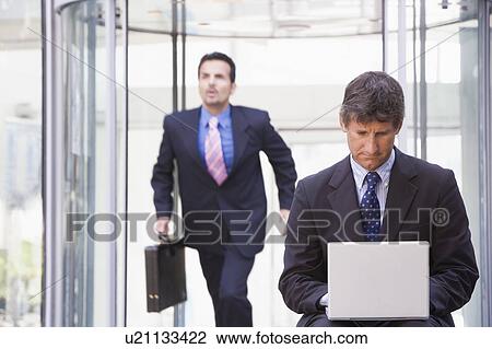 Stock Image - Businessman outdoors in front of building using laptop with businessman running in background (high key/selective focus). Fotosearch
