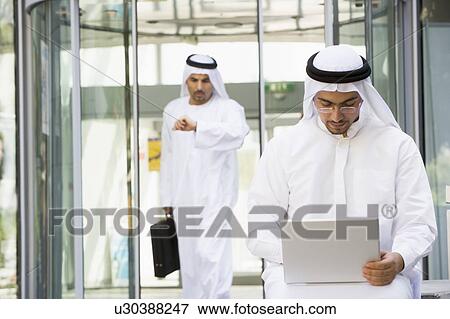 Stock Photo - Businessman outdoors using laptop with businessman walking in background checking watch (selective focus). Fotosearch