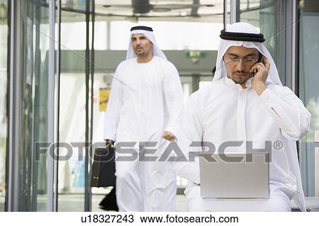 Stock Image - Businessman outdoors with laptop on cellular phone with businessman walking in background (selective focus). Fotosearch