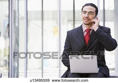Stock Photo - Businessman sitting outdoors by building with a laptop on cellular phone (high key/selective focus). Fotosearch