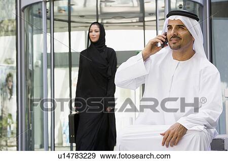 Stock Photo - Businessman sitting outdoors by building using cellular phone with businesswoman walking in background (selective focus). Fotosearch