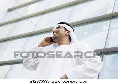 Stock Image - Businessman standing outdoors by building on cellular phone smiling (selective focus). Fotosearch