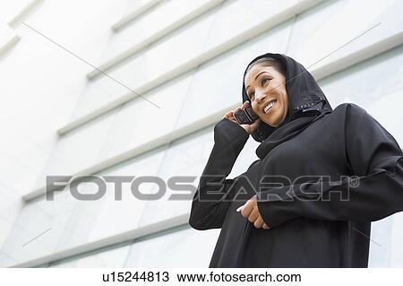 Stock Image - Businesswoman outdoors by building using cellular phone and smiling (selective focus). Fotosearch