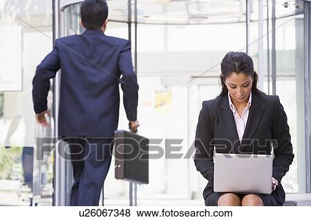 Businesswoman outdoors in front of building using laptop with businessman in background (high key/blur) View Large Photo Image Stock Photo - Businesswoman outdoors in front of building using laptop with businessman in background (high key/blur). Fotosearch
